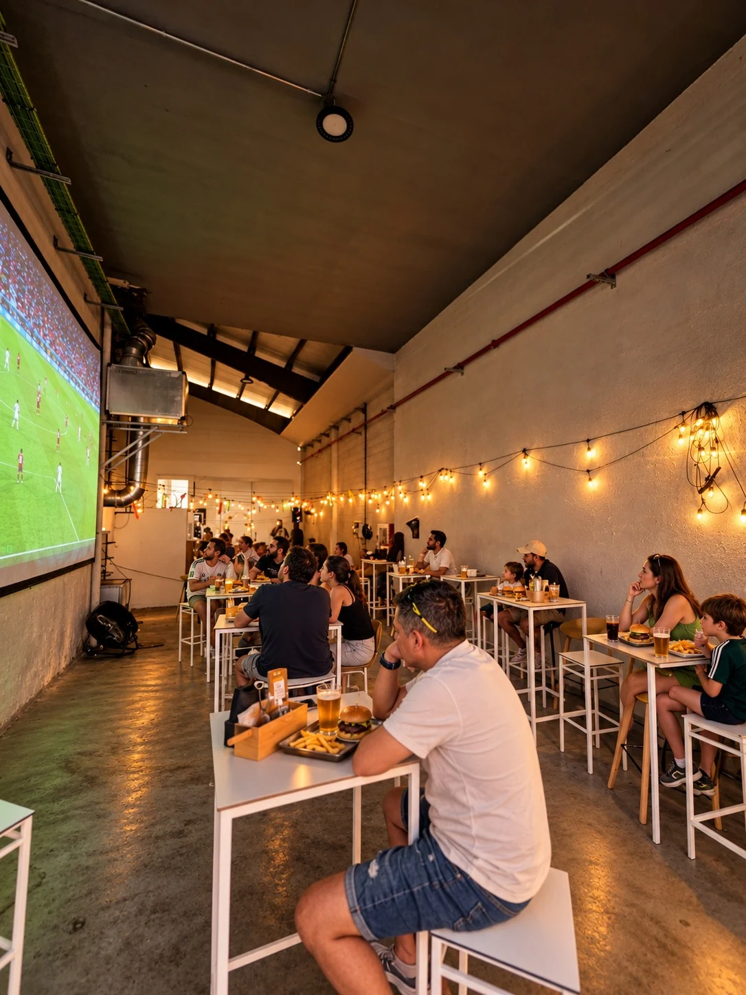 Ambientación de un partido de fútbol proyectado en la pantalla del local, con familias tomando burgers y bebidas